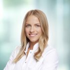 Uśmiechnięta kobieta w białym fartuchu lekarskim, stojąca na jasnym, rozmytym tle. Ma blond włosy i patrzy prosto w obiektyw.