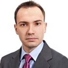 Mężczyzna spoglądający w obiektyw. Ubrany w ciemny garnitur i bordowy krawat