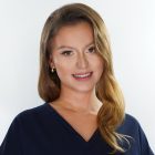 Uśmiechnięta kobieta w granatowym uniformie medycznym pozuje na jasnym tle – ma długie, falowane włosy i delikatny makijaż.