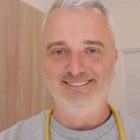 Uśmiechnięty mężczyzna z siwymi włosami i zarostem, w szarej bluzie, z żółtym stetoskopem na szyi, stojący na tle jasnego pomieszczenia