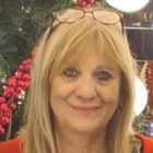 Uśmiechnięta kobieta z blond włosami, w okularach założonych na głowę, stoi na tle świątecznego drzewka z czerwonymi dekoracjami